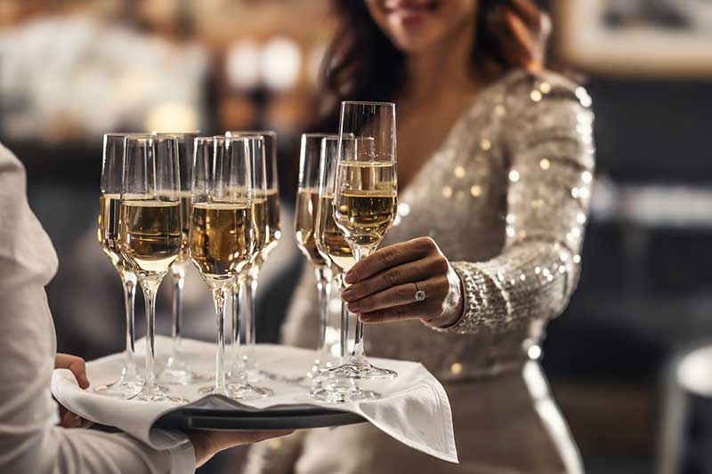 a holiday party venue in quincy ma - photo of waiter serving glasses of champagne; a woman dressed for a party reaches for one of the glasses from the waiter's tray